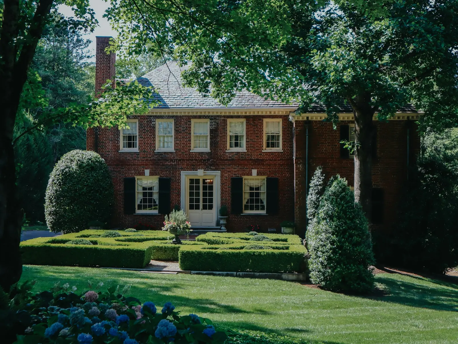 A brick home framed by mature trees, clipped hedges, and a green lawn