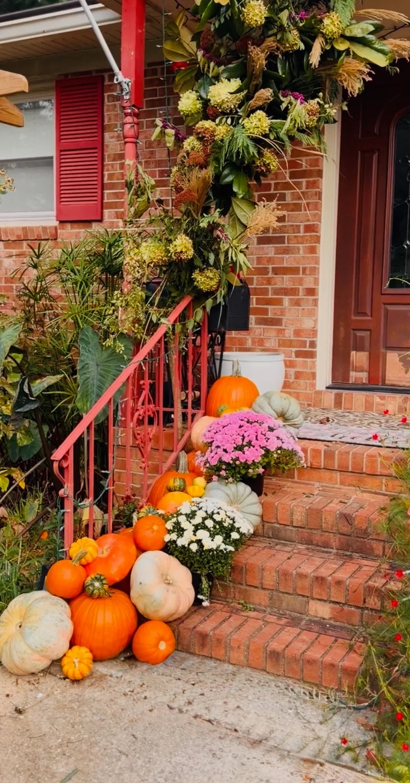 Pumpkins and pink and white mums on brick steps beneath a leafy fall arch